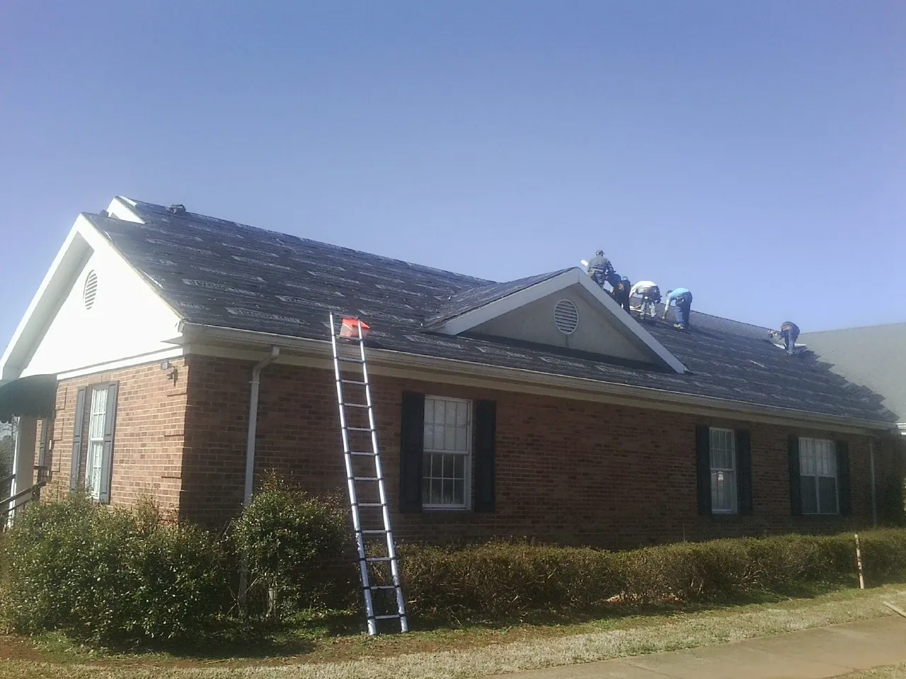Crew tearing shingles off a brick ranch roof with a ladder set — Metal Roof Repair work by our crew in Gas City