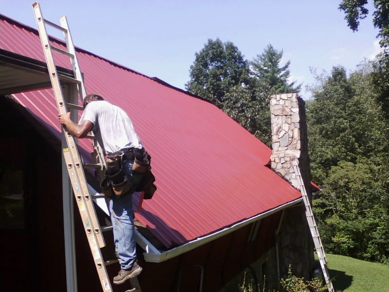 Roofer on a ladder beside a red metal roof and stone chimney — Soffit & Fascia Repair by Roofing Brothers in Gas City